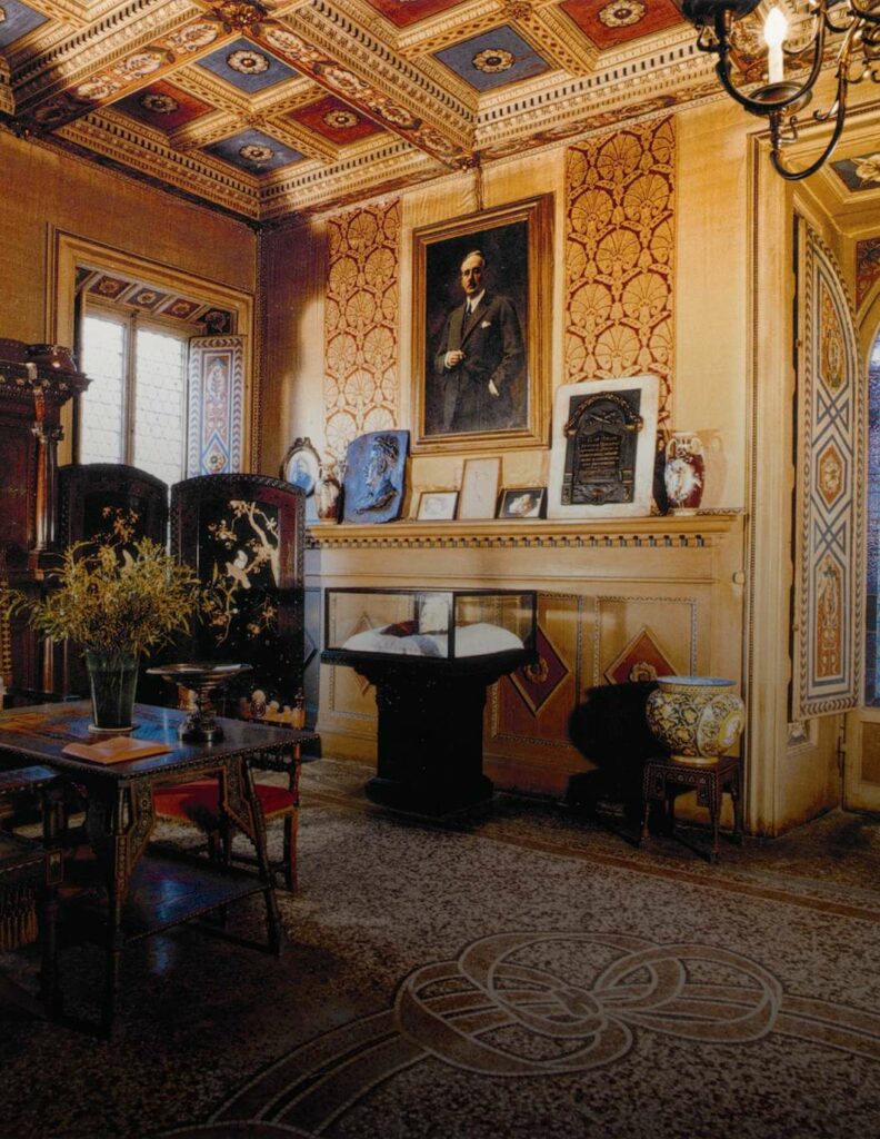 Vista parziale del salone di Villa Puccini. Il soffitto a cassettoni è una delle caratteristiche principali della stanza, con l'alternanza dei colori blu e rosso per ogni riquadro. Sulle pareti, una carta da pareti damascata con motivi orientali, lo stesso stile è richiamato per il paravento. Un quadro raffigurante il Maestro è appeso al muro, mentre al centro della stanza si trova un tavolo in legno scuro. Sotto il quadro, una teca con la maschera funerea di Puccini. Il pavimento in marmo presenta decorazioni tipiche dell'epoca, con fiocchi avvolti su loro stessi che si stagliano sulla superficie.