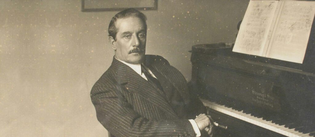 Foto d'epoca che ritrae il maestro in una posa di 3:4 con il volto rivolto verso l'obiettivo. È seduto accanto a un pianoforte, vestito con un elegante abito gessato.