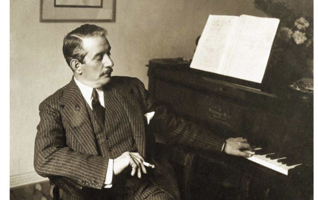 Foto d'epoca che ritrae il maestro accanto a un pianoforte, vestito con un elegante abito gessato.
