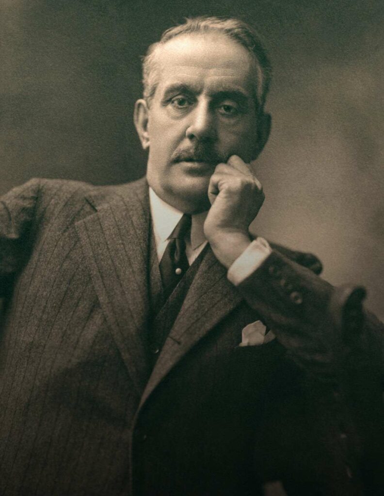 Foto d'epoca dell'ultimo Puccini, in cui lo si vede seduto mentre guarda in camera e poggia la testa sulla mano. Nell'immagine, il celebre compositore indossa un elegante abito gessato.