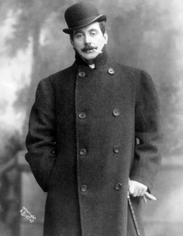 Giacomo Puccini indossa un elegante cappotto invernale, una bombetta e impugna il suo bastone. Tra le labbra, tiene una sigaretta.