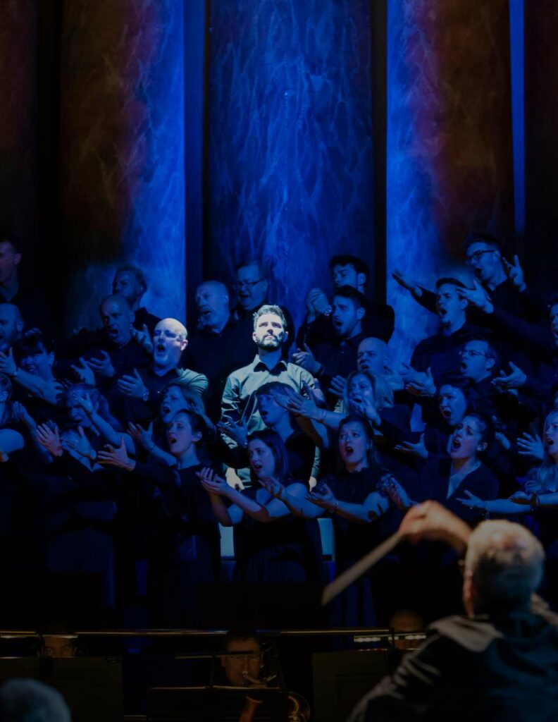 Scena dell'opera Turandot al Leeds Town Hall. Il tenore è illuminato da una potente luce bianca, mentre il coro, rimasto in ombra, affolla il palco.